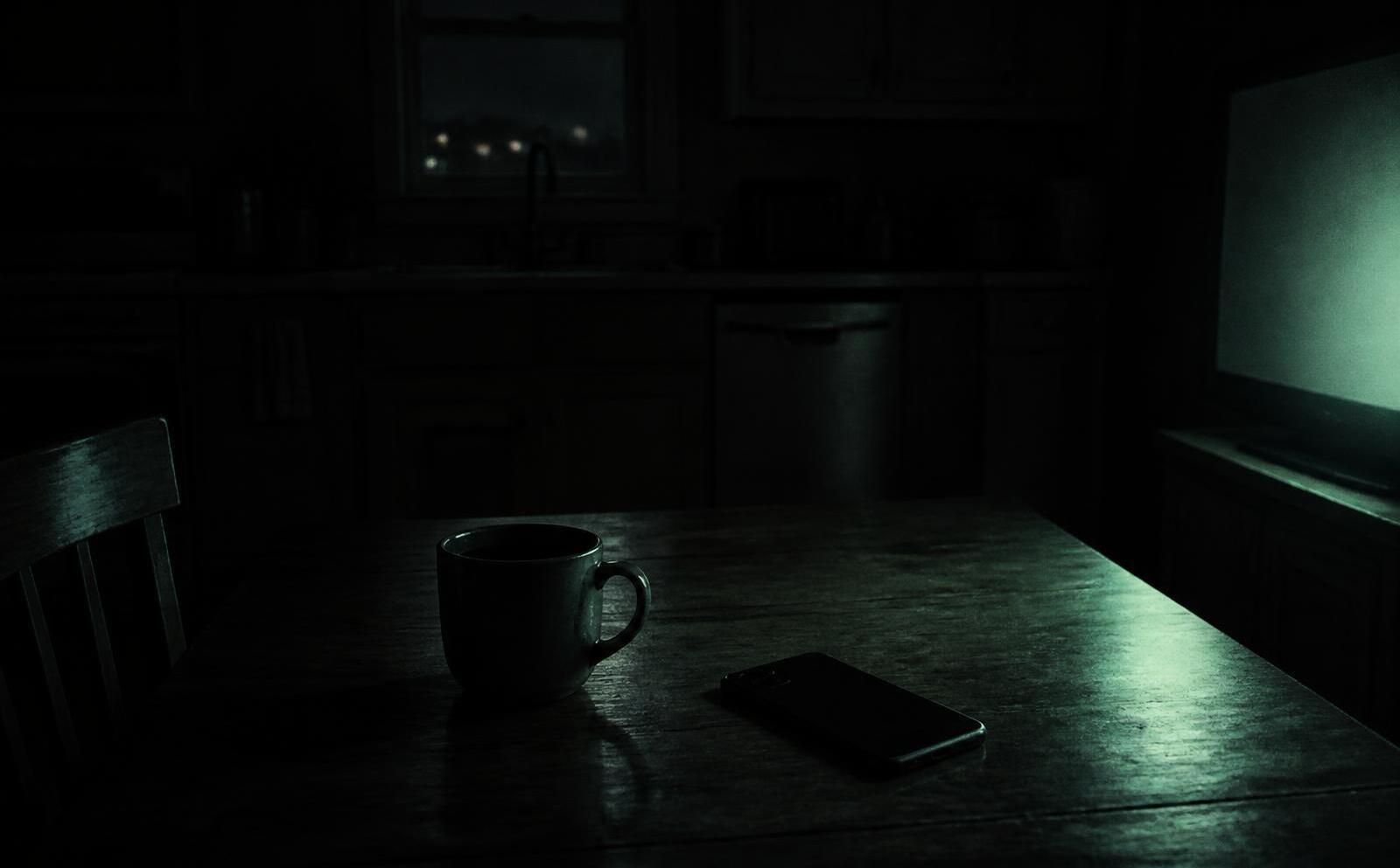 A dark kitchen table at night, a phone face down beside a cold mug